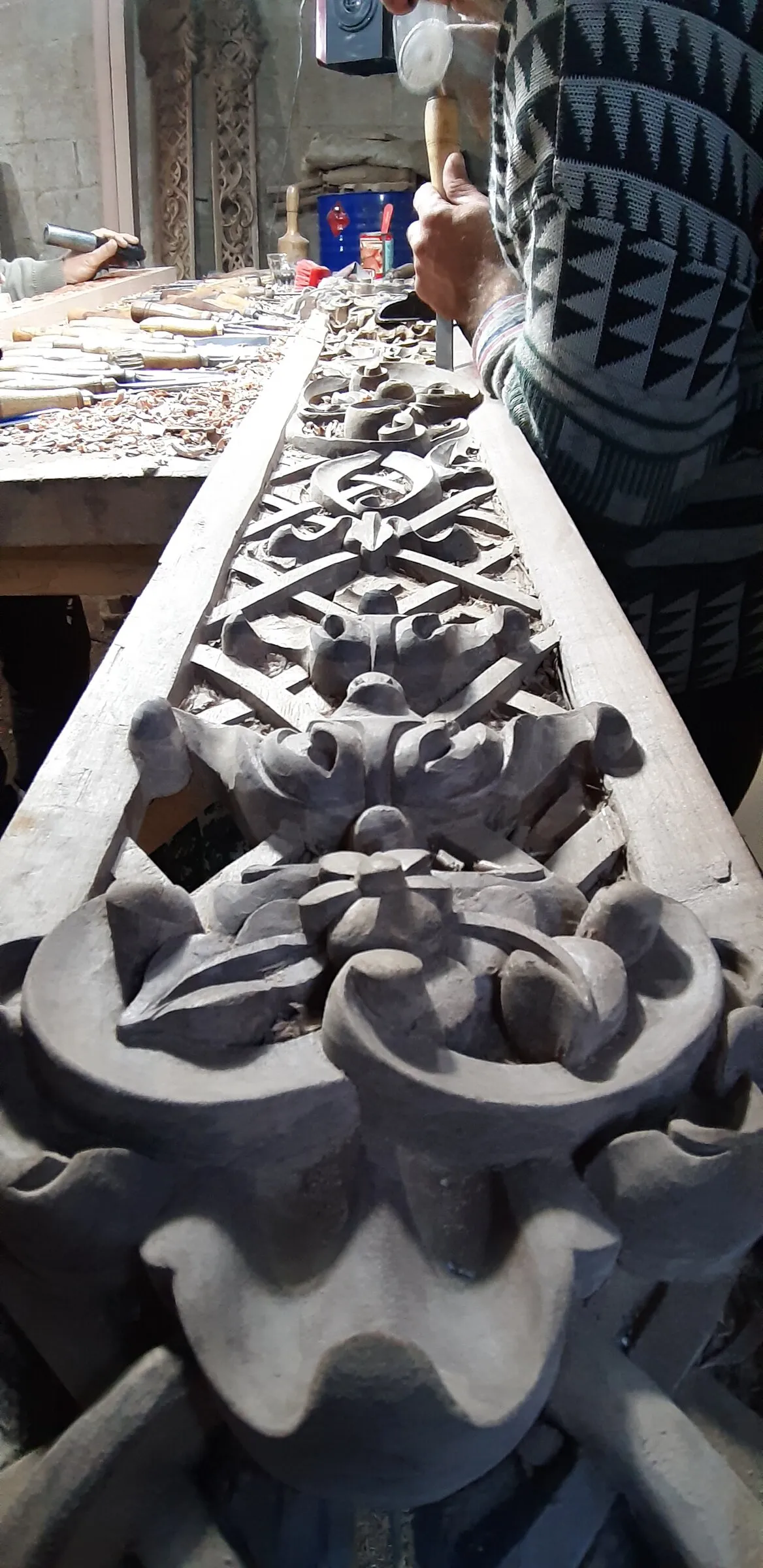 A long decorative carving lies across a bench as workers shape its surface.