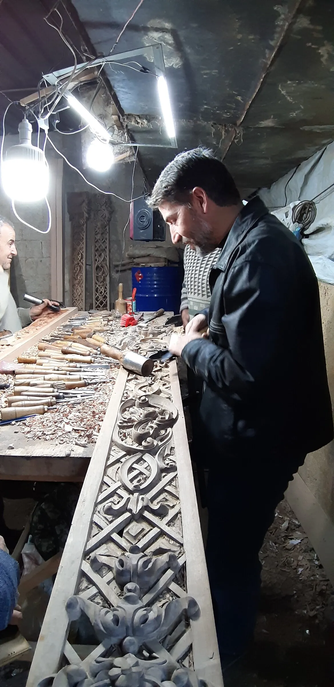 A craftsperson works beside a long ornamental carving under bright workshop lights.