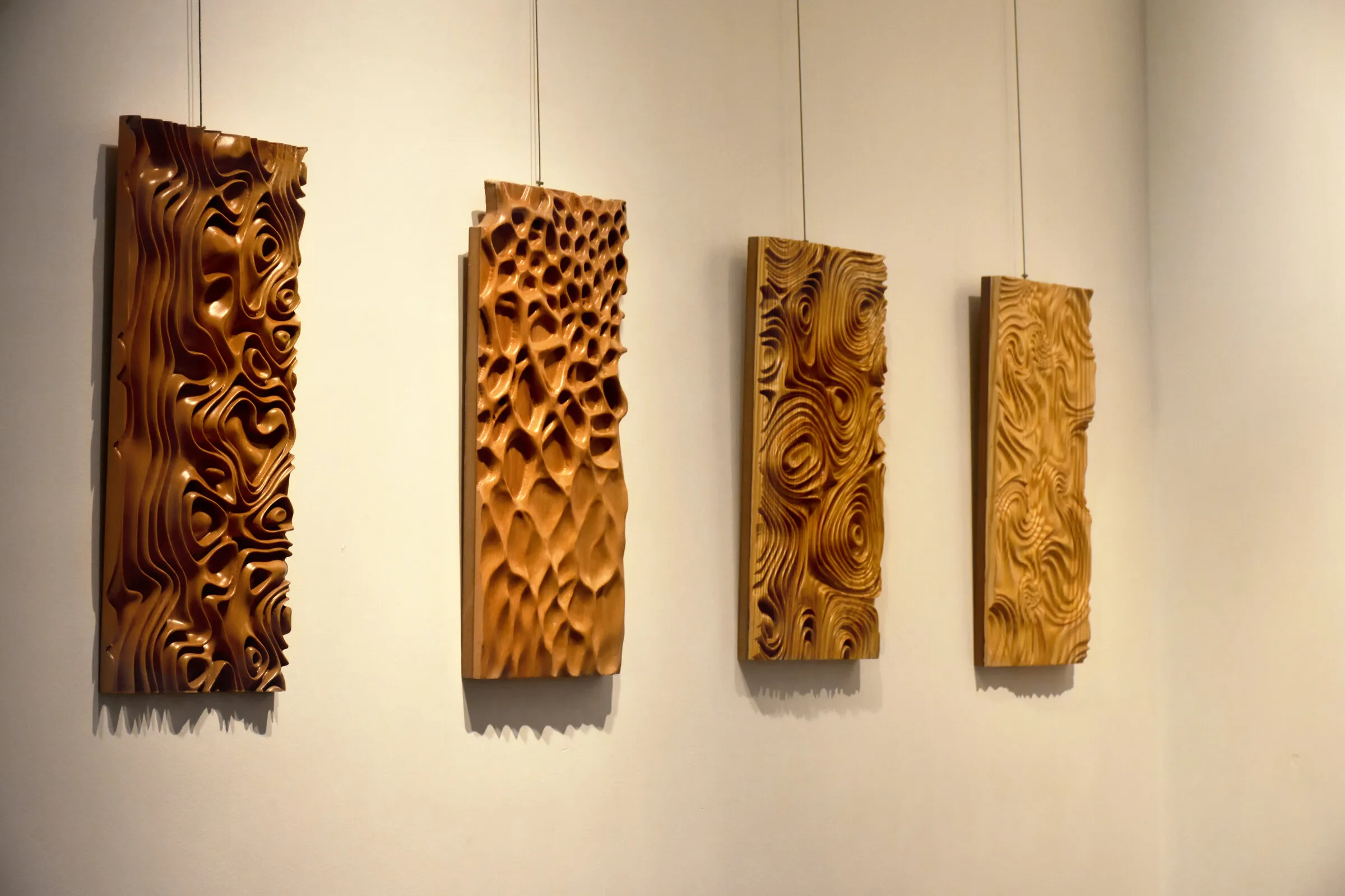 Four carved wooden panels hang in a row on a pale gallery wall at a design exhibition.
